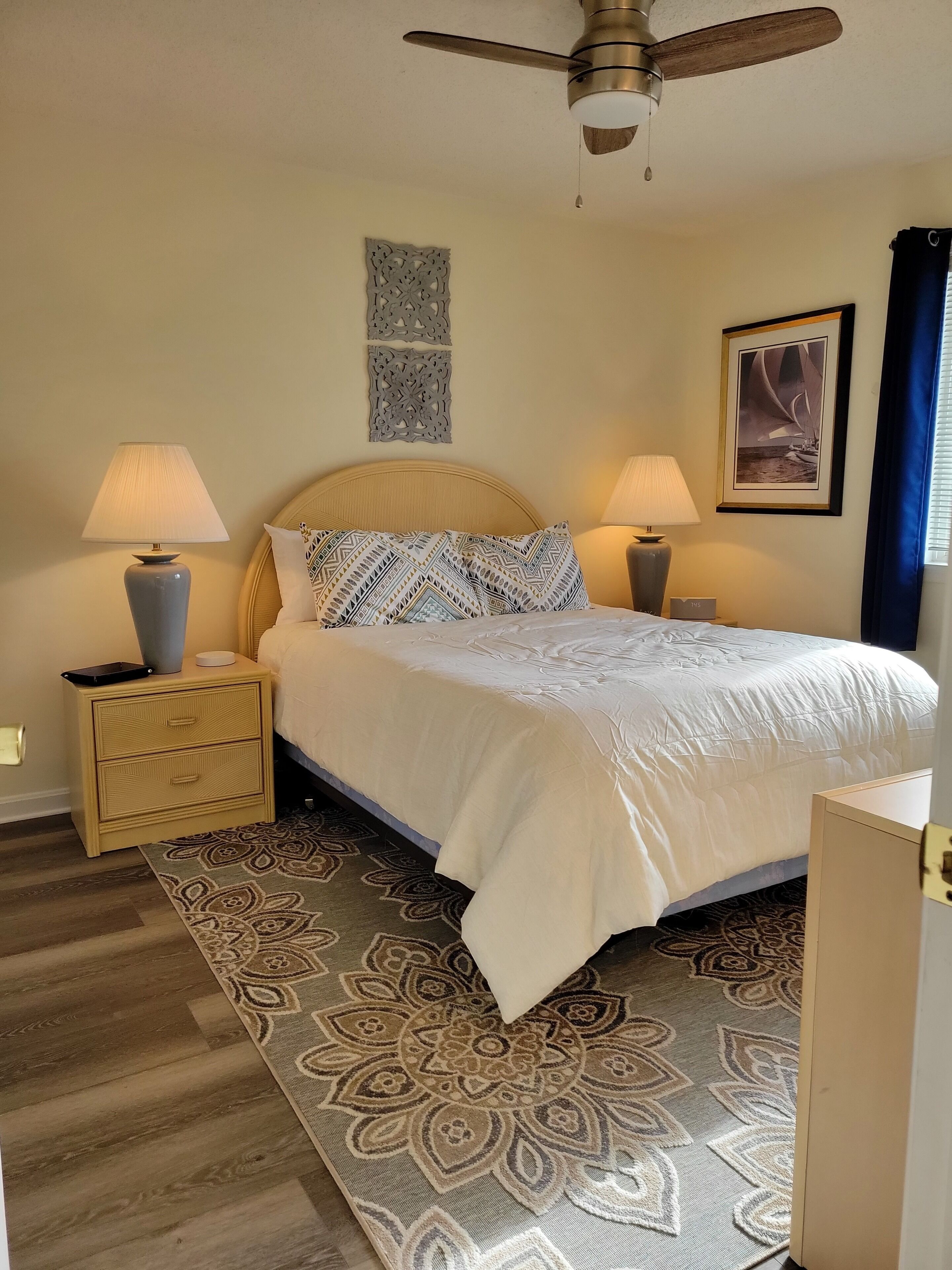 A bedroom with a bed, two bedside tables with lamps, a ceiling fan, a patterned rug, and a wall-mounted artwork.