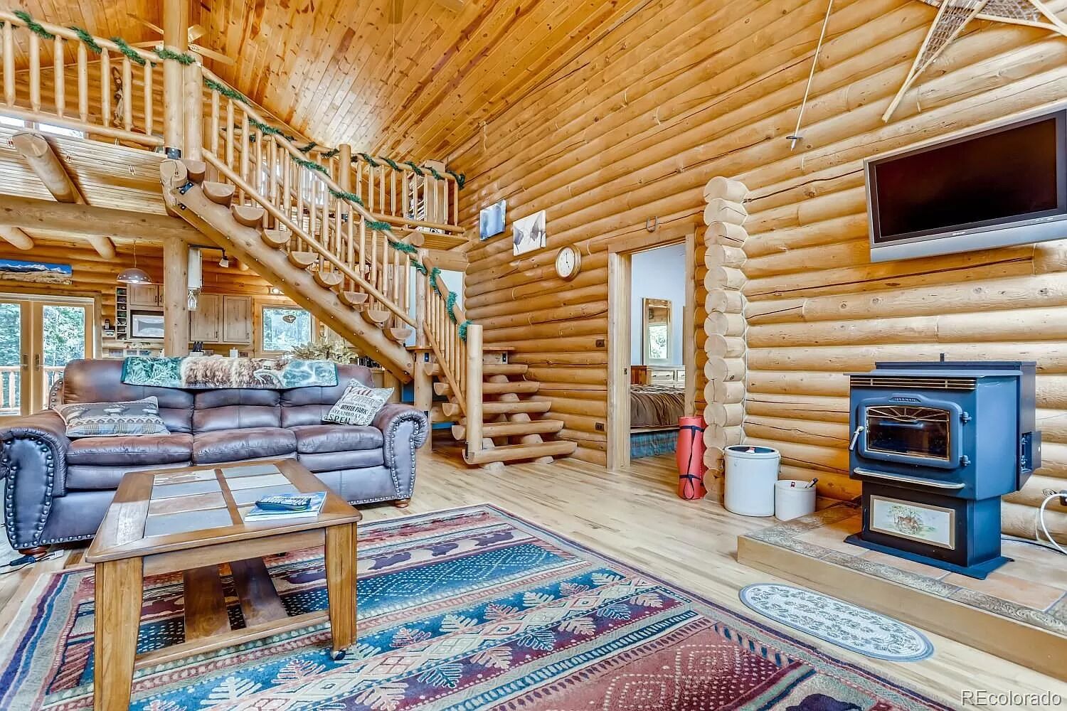 A log cabin interior with a staircase, a sofa, a TV, and a wood stove.