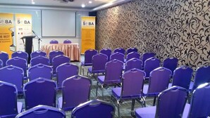 Sala de conferências com fileiras de cadeiras roxas, um púlpito e uma tela de projeção.
