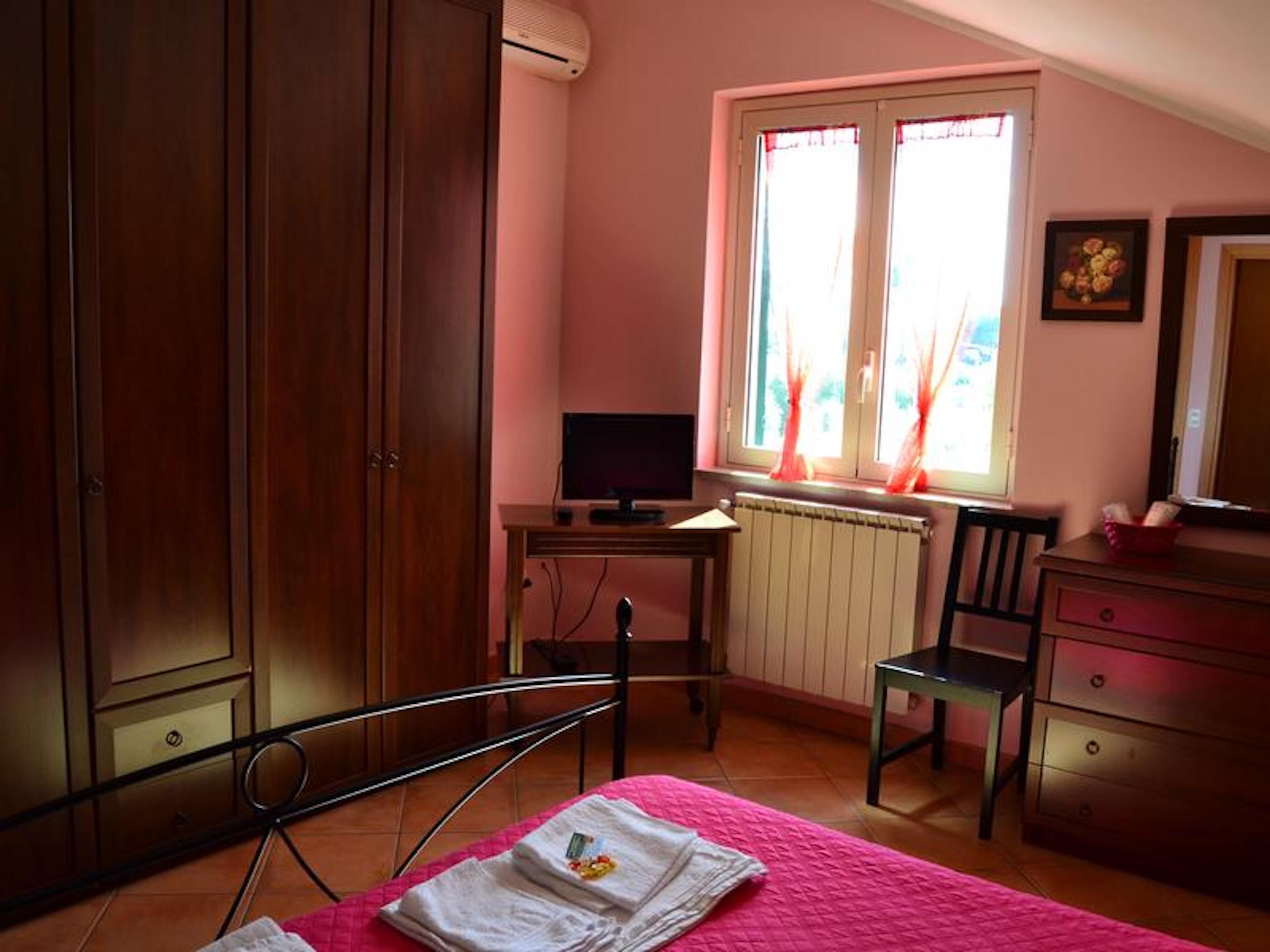 A bedroom with a bed, a desk with a computer, a chair, a wardrobe, and a window with curtains.