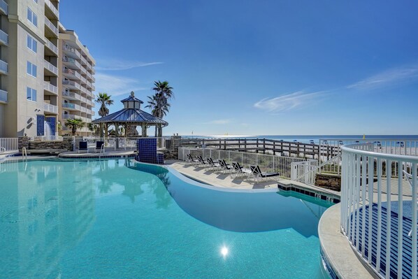 A swimming pool with lounge chairs and a gazebo, surrounded by a fence, with a building and beach in the background.