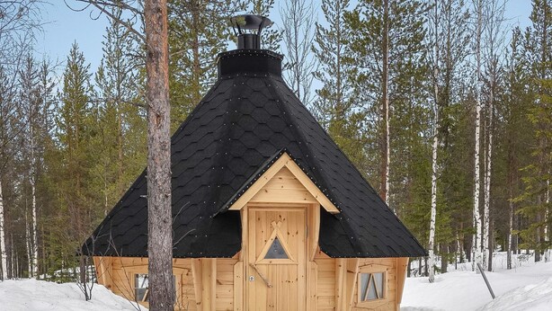 A wooden cabin with a black roof, surrounded by snow and trees.