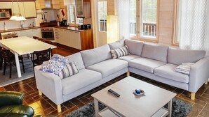 A modern living room with a sectional sofa, a dining table, and a kitchen area with wooden cabinets and a chandelier.