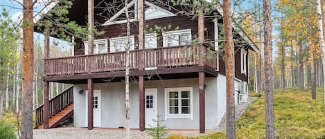 A two-story cabin with a wooden deck and a covered porch, surrounded by trees and a forested landscape.