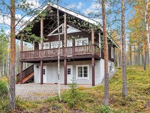 A two-story cabin with a wooden deck and a covered porch, surrounded by trees and a forested landscape.