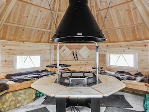 A wooden interior with a central black conical structure, possibly a fireplace, surrounded by a metal frame. There are two windows with white frames and a view of the outside. Fur rugs and a bench are visible on the floor.