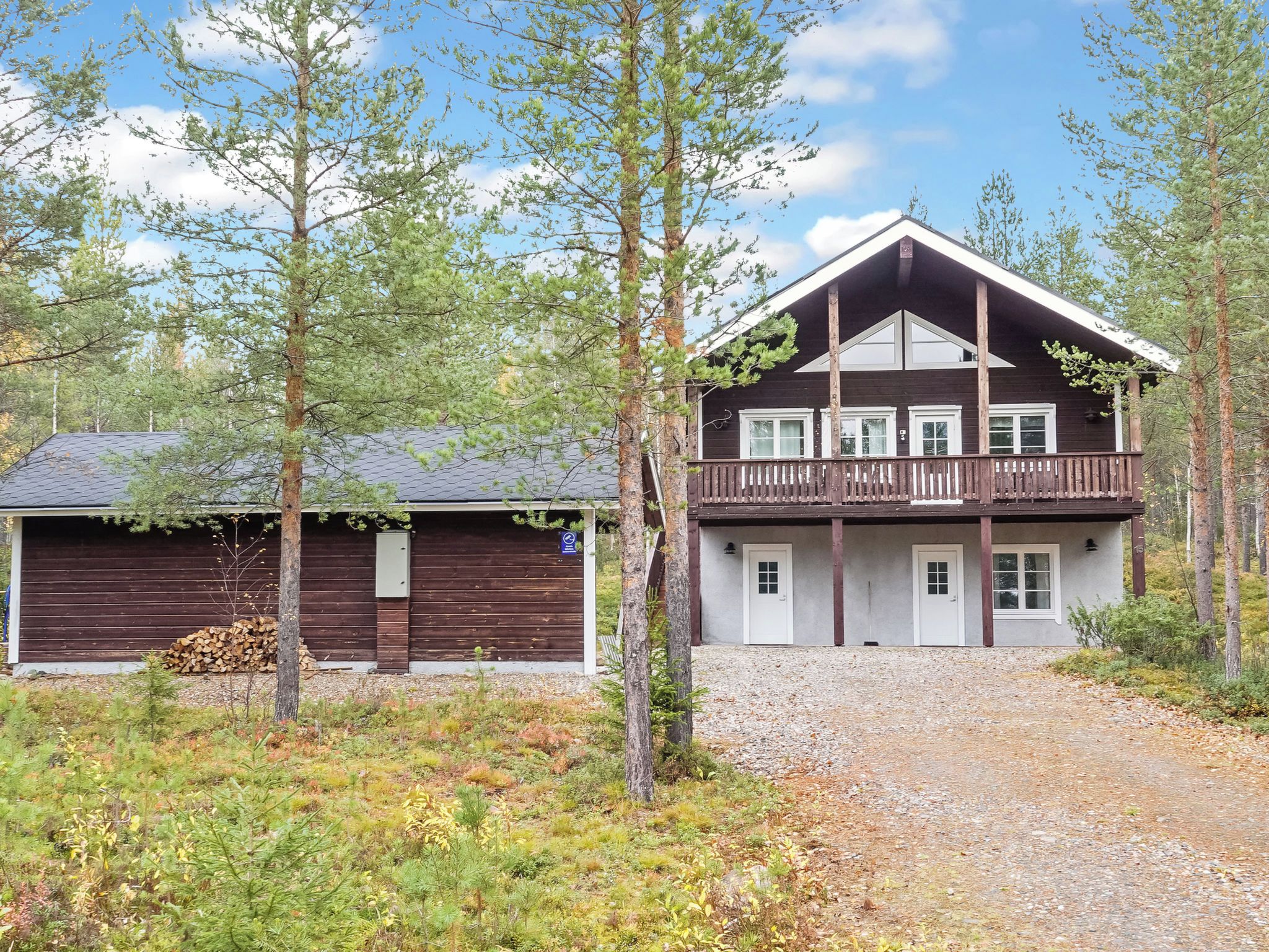 A two-story wooden cabin with a balcony, surrounded by trees and a gravel driveway.