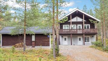 A two-story wooden cabin with a balcony, surrounded by trees and a gravel driveway.