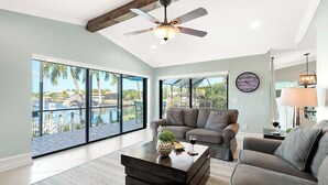 Ein geräumiges Wohnzimmer mit großem Fenster und Blick auf einen Pool und Palmen, einem Deckenventilator und einer Wanduhr.