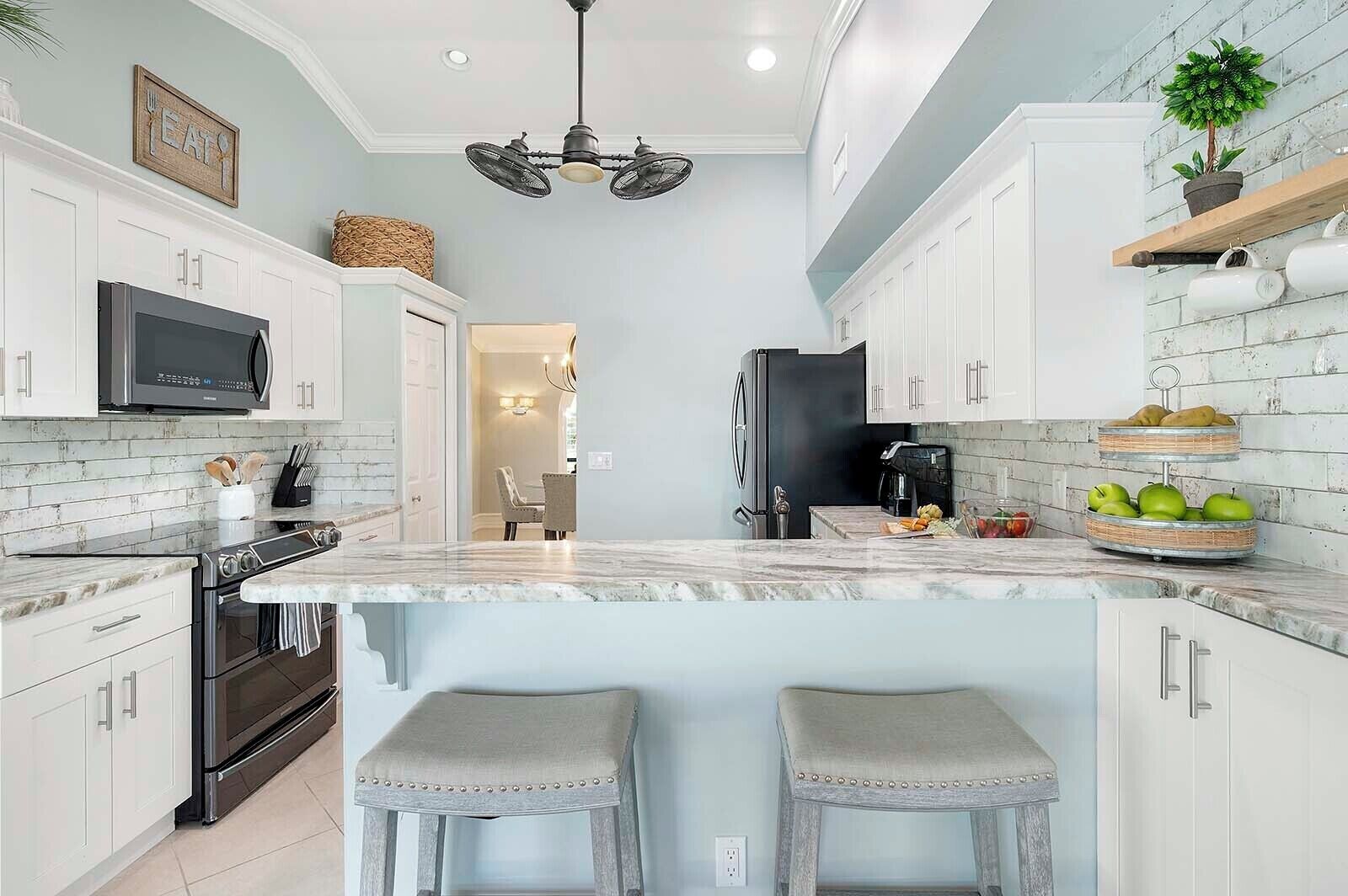 Cocina moderna con armarios blancos, un refrigerador negro, microondas y encimera de mármol. Se observan dos taburetes de bar y una zona de comedor al fondo.