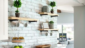 Eine Küche mit weiß gefliester Spritzschutzwand, Holzregalen mit Tassen und Pflanzen sowie einer Schüssel grüner Äpfel auf der Arbeitsfläche.