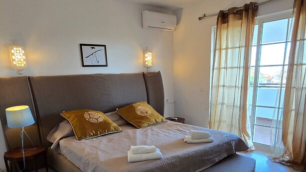 A modern bedroom with a large bed, a bedside table, an air conditioning unit, and a window with curtains.