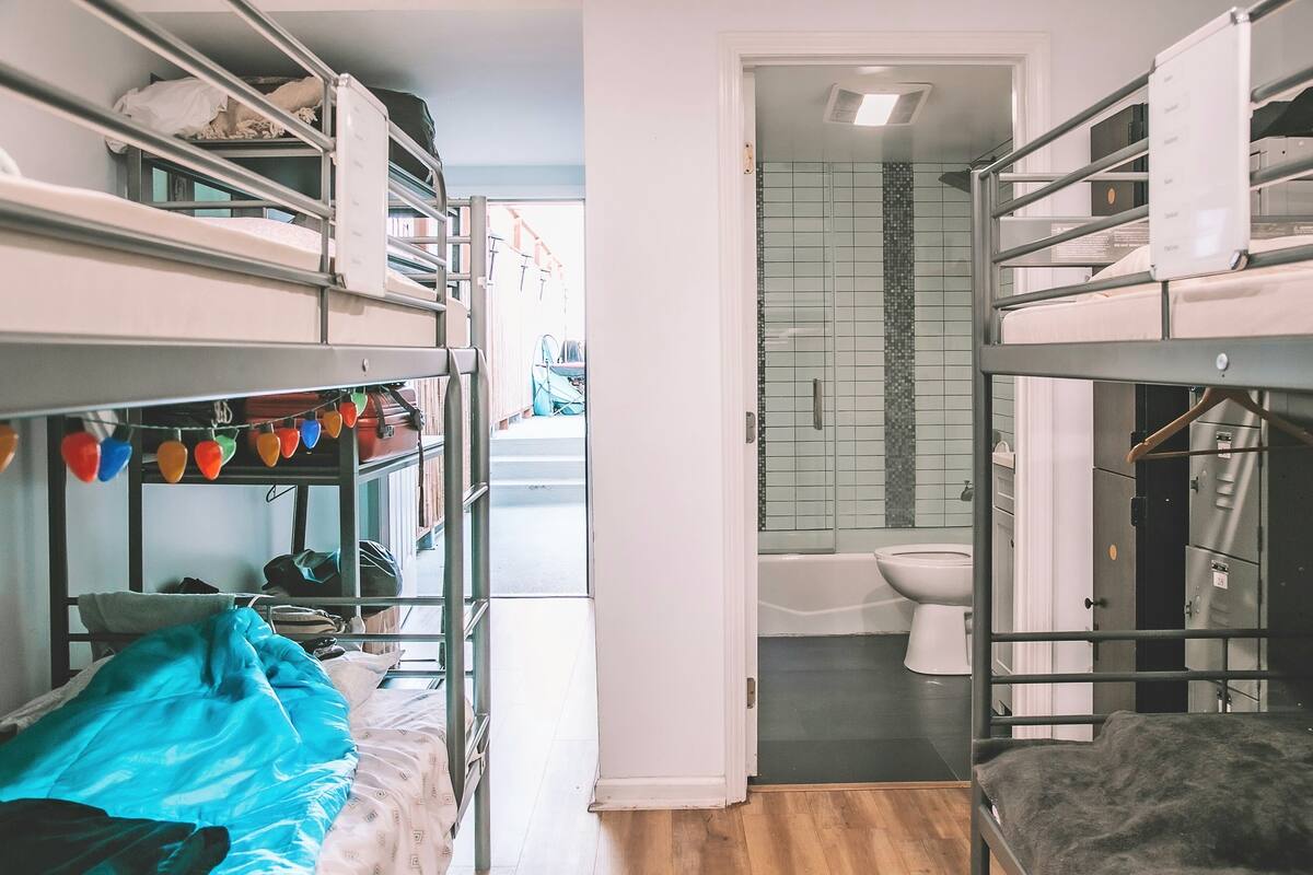 A dormitory room with bunk beds, a bathroom with a toilet and shower, and a view of the street outside.