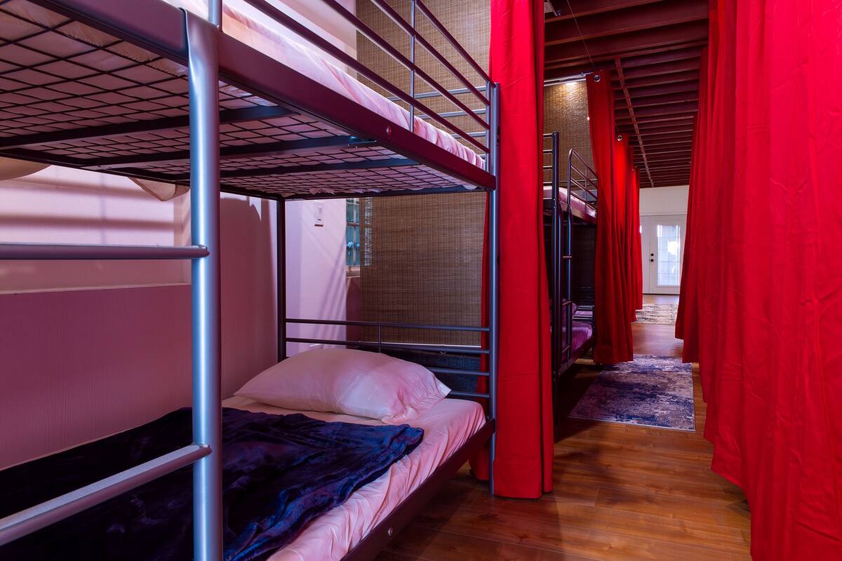 A bunk bed room with a red curtain, wooden flooring, and a view into another room.
