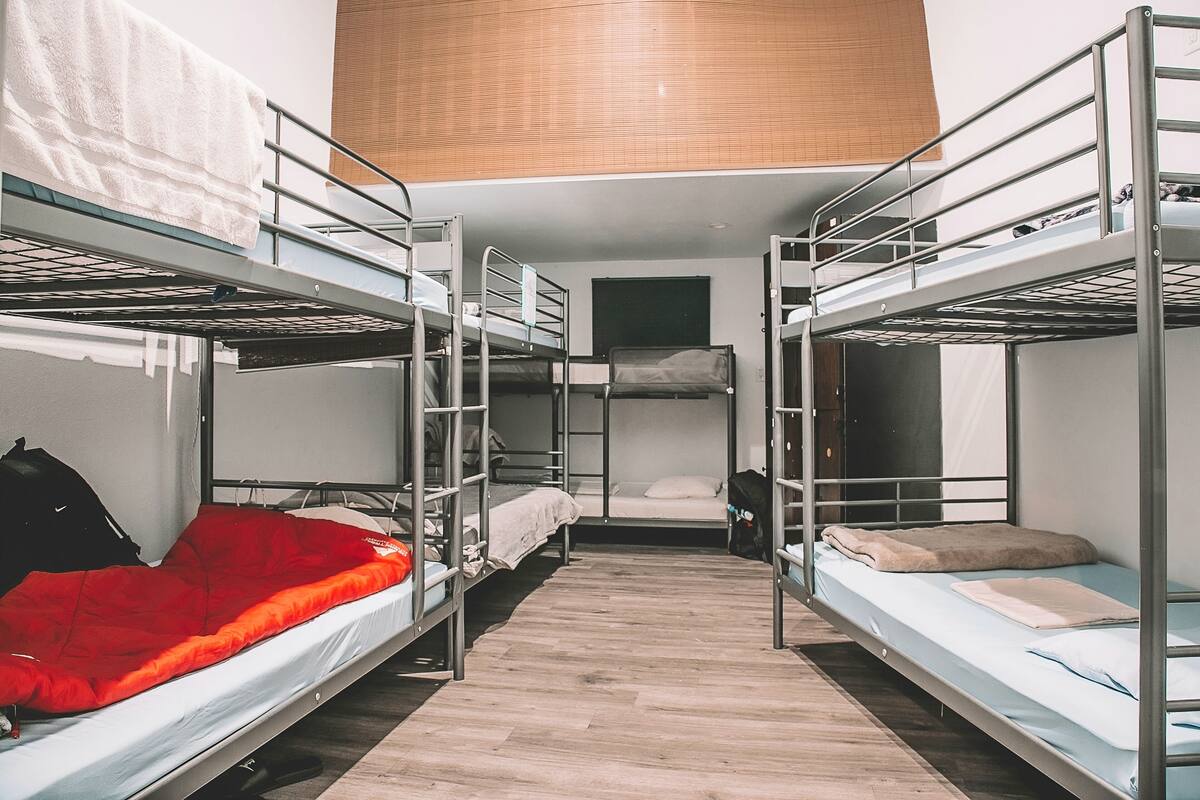 A dormitory room with bunk beds, a wooden floor, and a window with blinds.