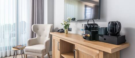 A modern hotel room with a wooden console table, a beige armchair, a flat-screen TV, and a coffee maker.