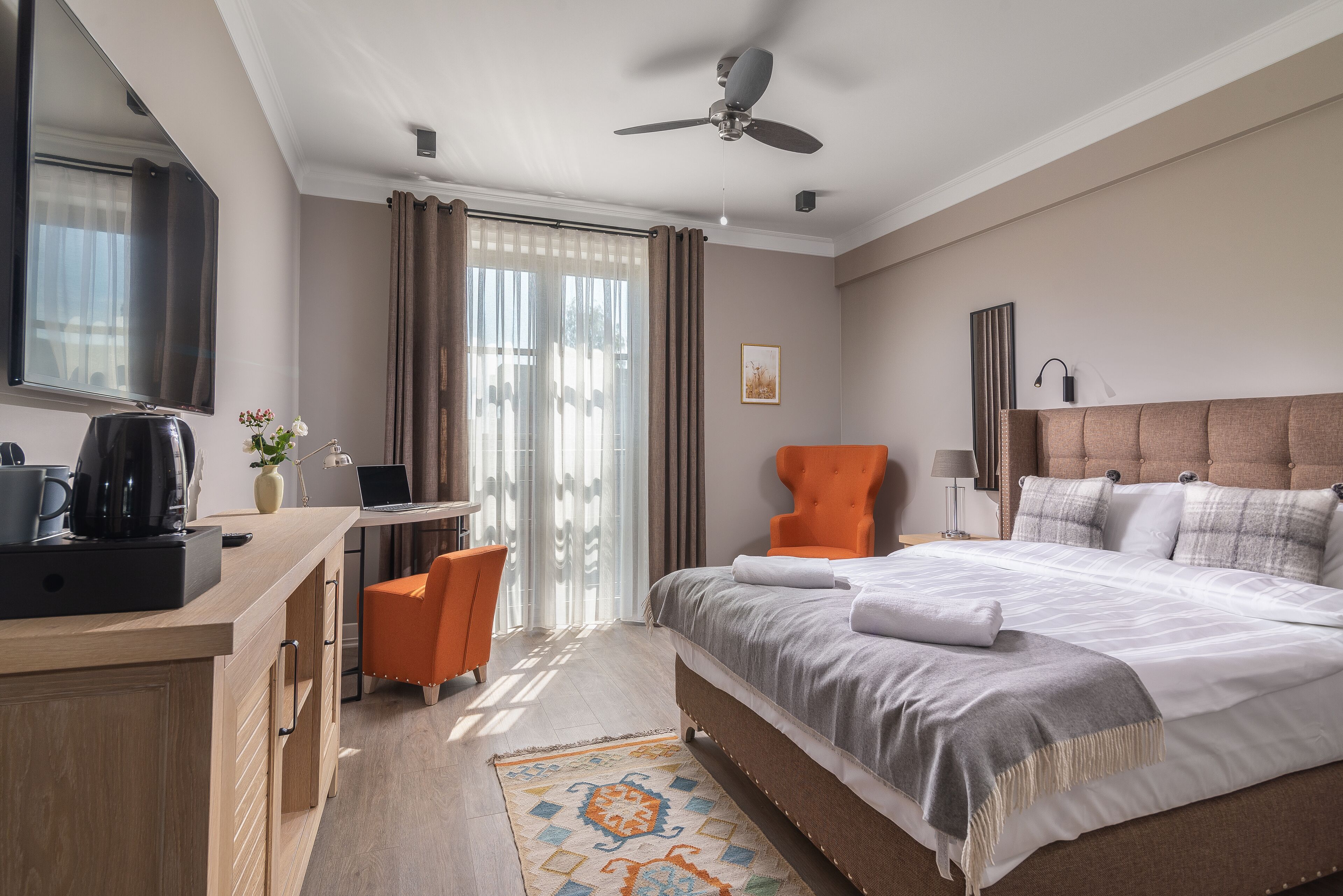 A hotel room with a bed, a desk, an orange chair, a television, and a ceiling fan.
