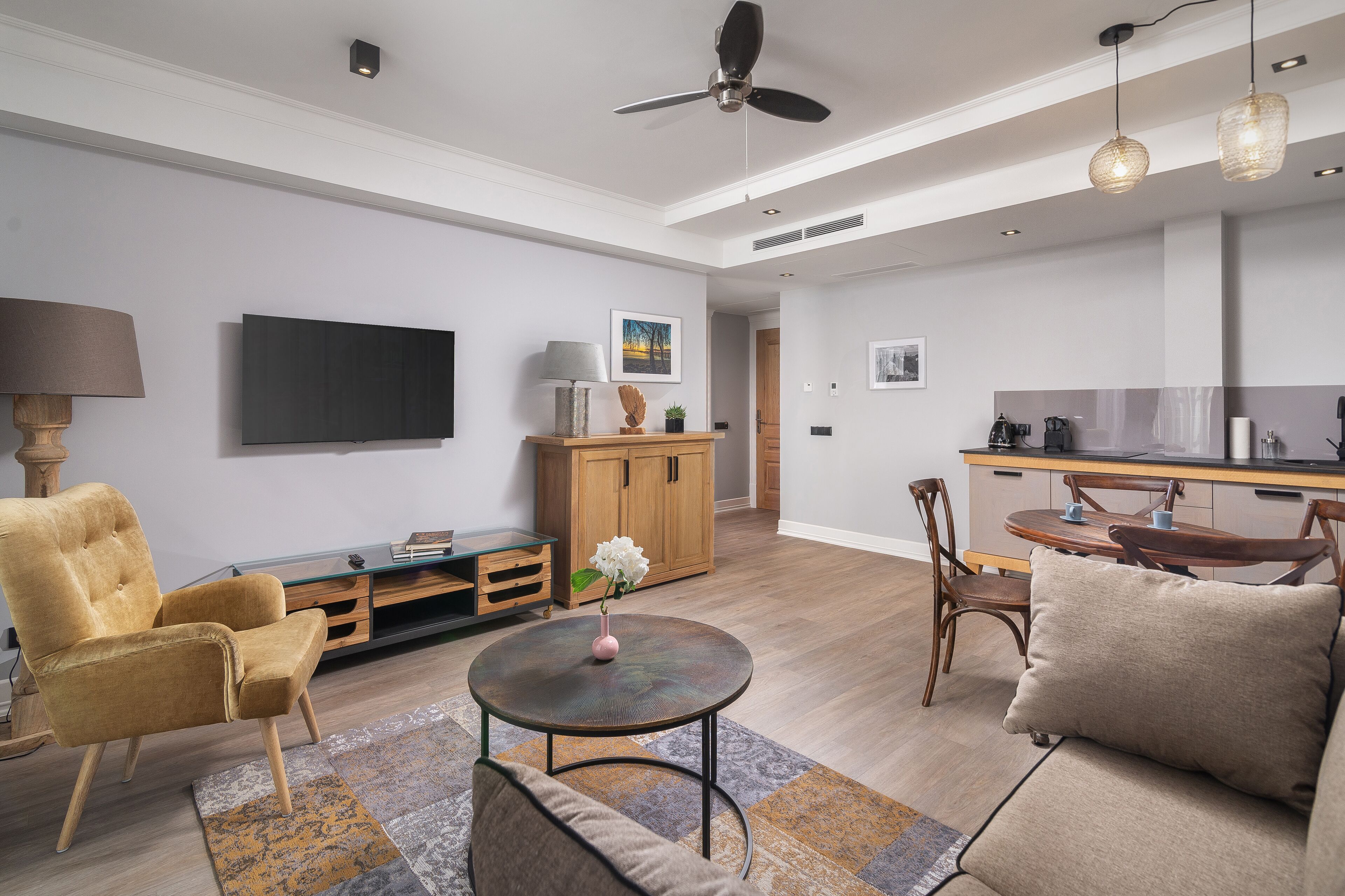Un salon moderne avec un canapé, un fauteuil et une table basse ronde. Un téléviseur est fixé au mur, et il y a également un meuble en bois.