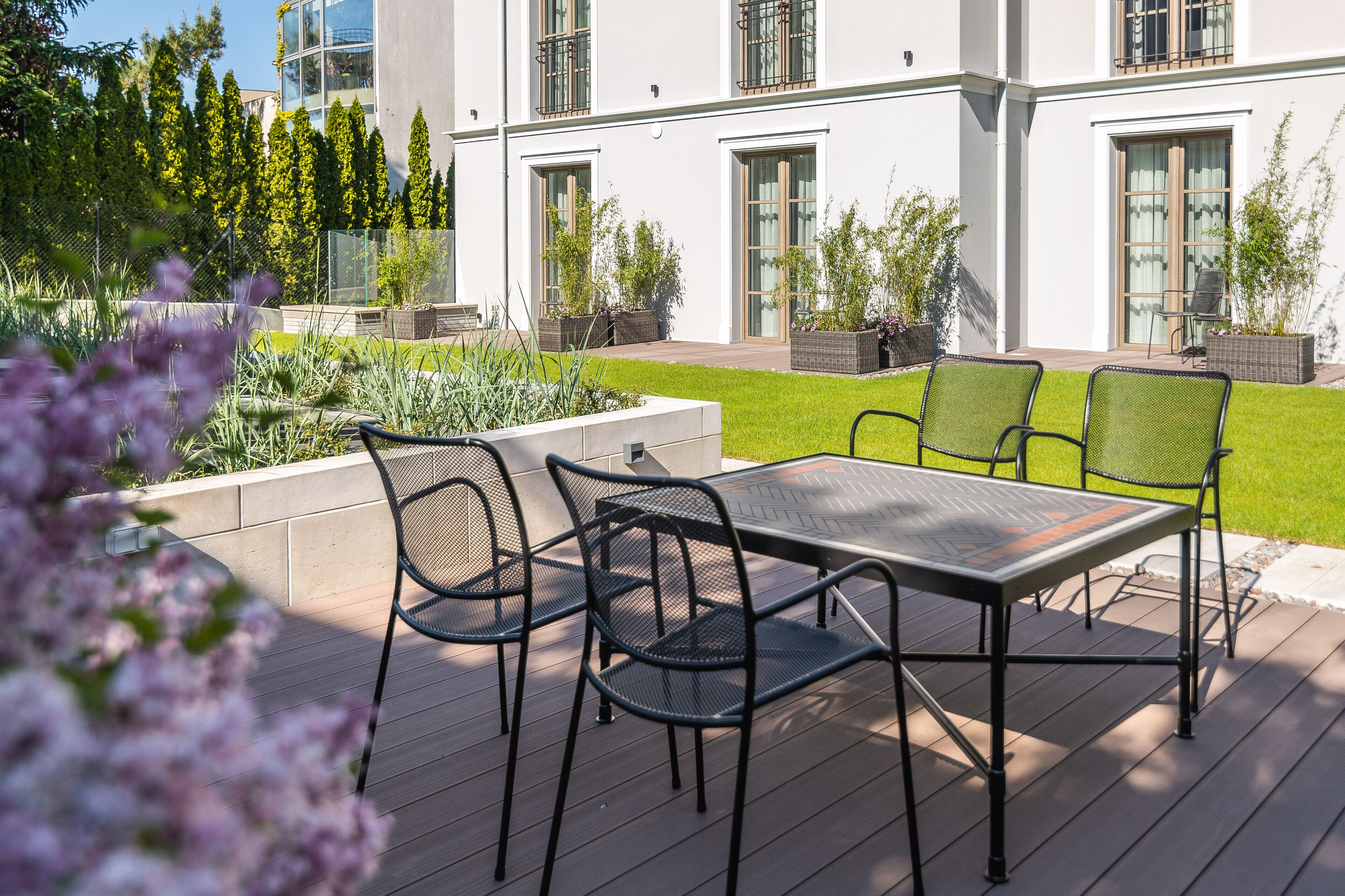 Une terrasse avec une table et des chaises, entourée d’un jardin où poussent différentes plantes et délimitée par une clôture.