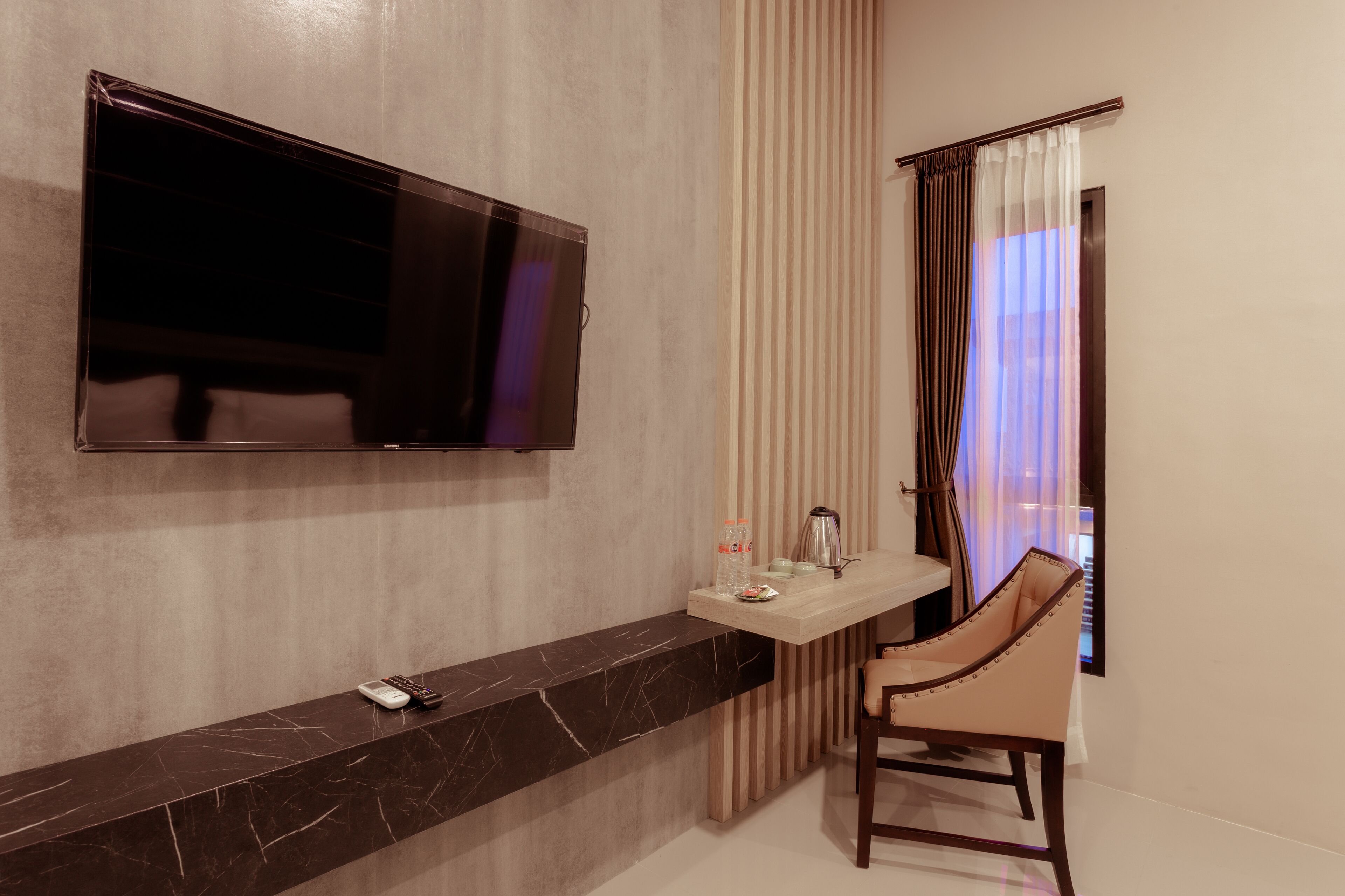 A modern hotel room with a large flat-screen TV mounted on the wall, a marble console table, a chair, and a window with curtains.