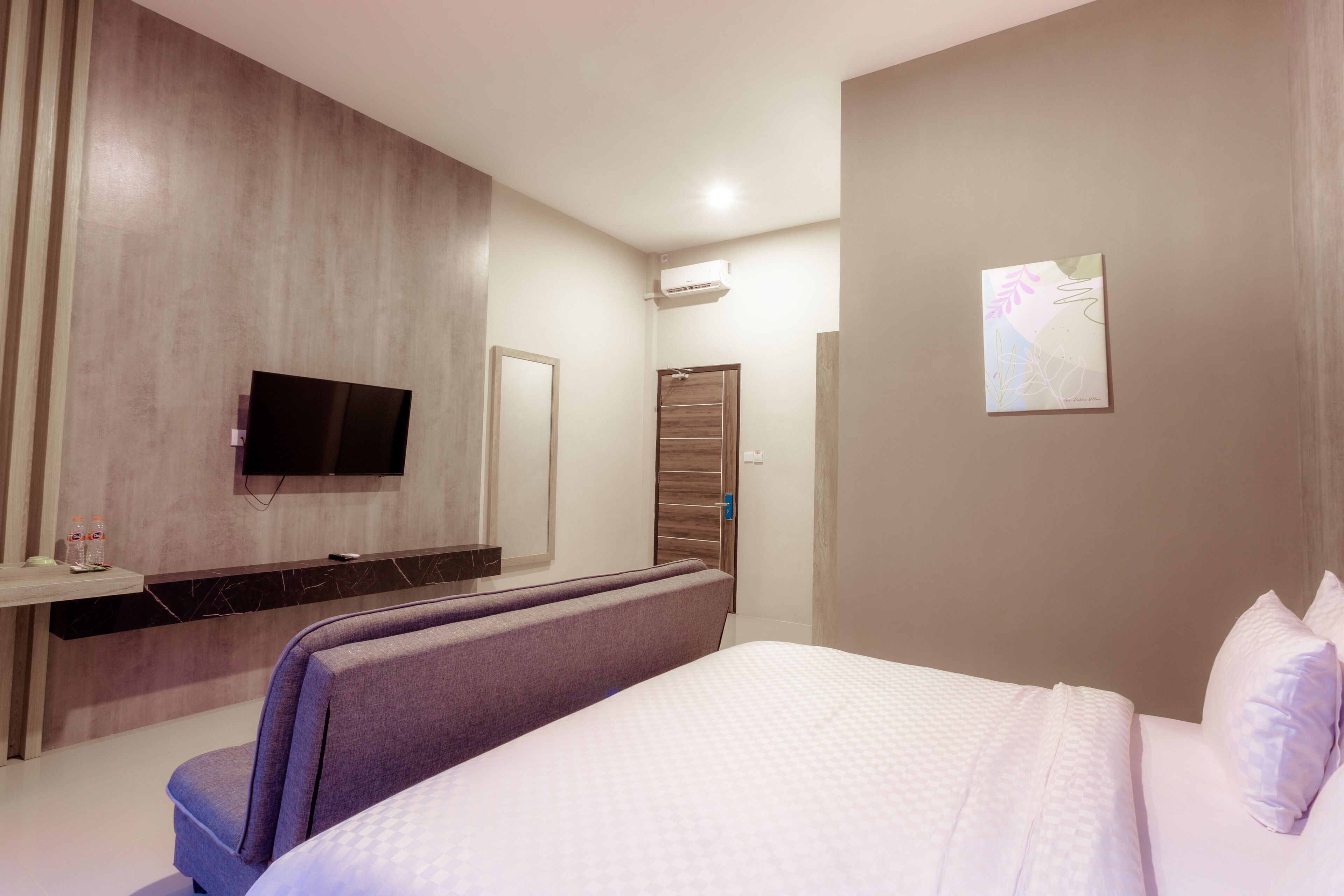 A modern hotel room with a large bed, a flat-screen TV mounted on the wall, a small desk with a mirror, and a wall-mounted air conditioning unit.