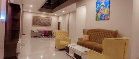 A modern living room with a brown sofa, yellow armchairs, and a white coffee table. There is a painting on the wall and a staircase in the background.