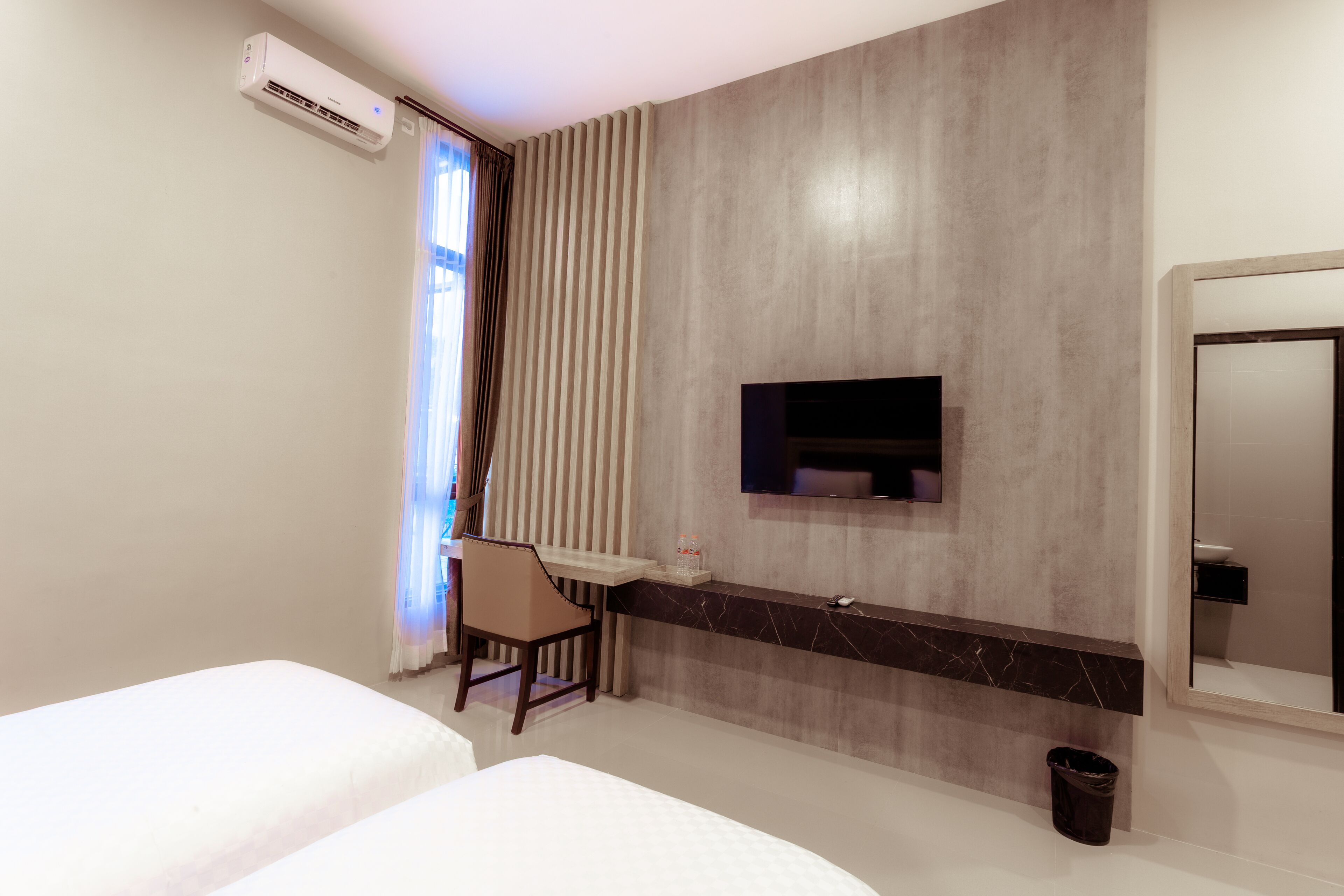 A modern hotel room with a bed, a desk, a chair, a TV mounted on the wall, and a bathroom mirror.