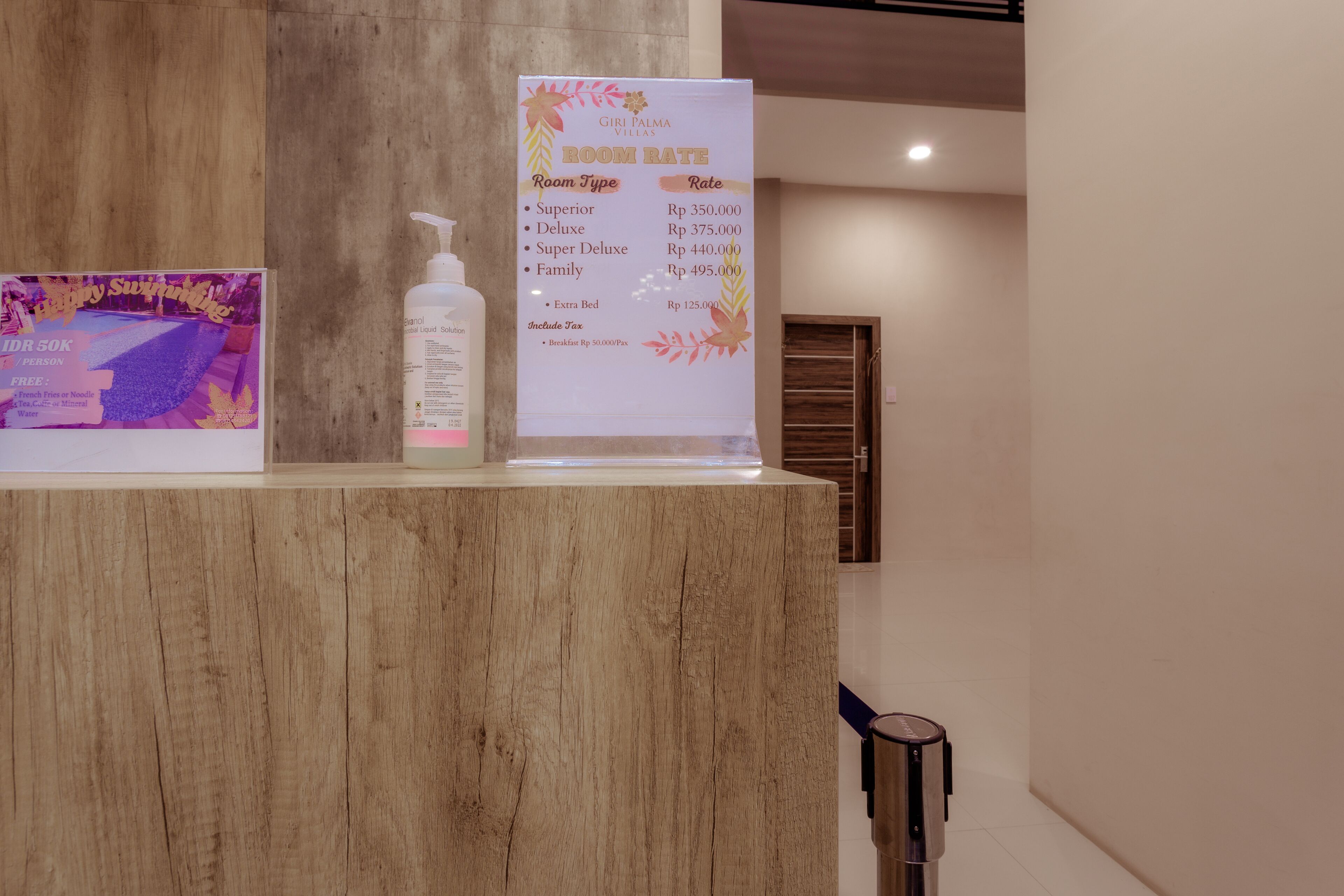 A hotel reception desk with a wooden counter, a room rate sign, a bottle of lotion, and a promotional poster.