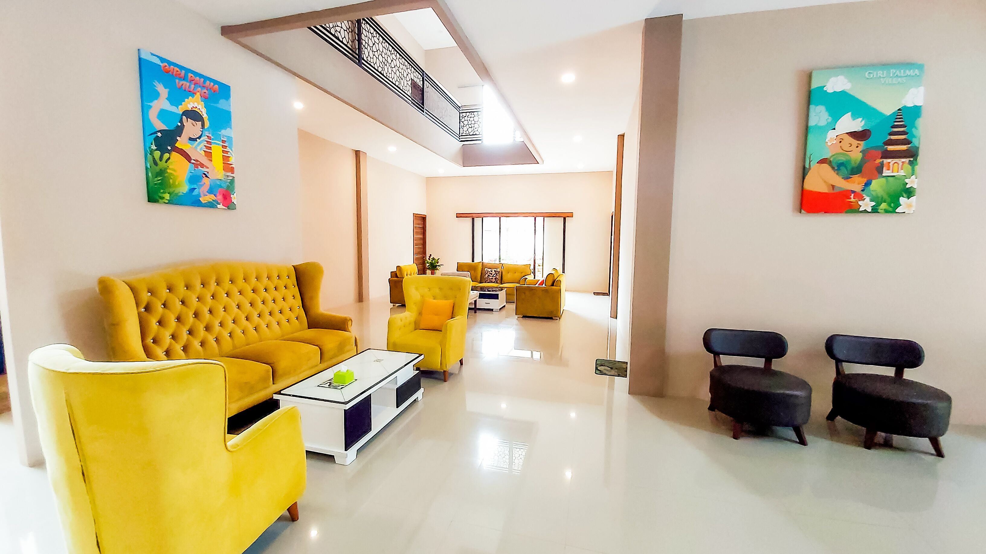Una sala moderna con un sofá amarillo, una mesa de centro y dos sillones. Hay dos cuadros en las paredes, uno con una escena tropical y otro con una escena cultural.