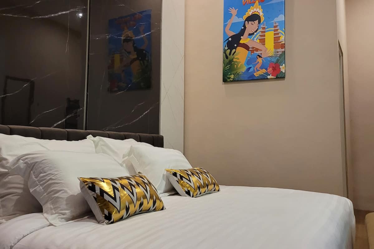 Ett hotellrum med en stor säng, vita sängkläder och två dekorativa kuddar. På väggen hänger en tavla som föreställer ett tropiskt landskap.