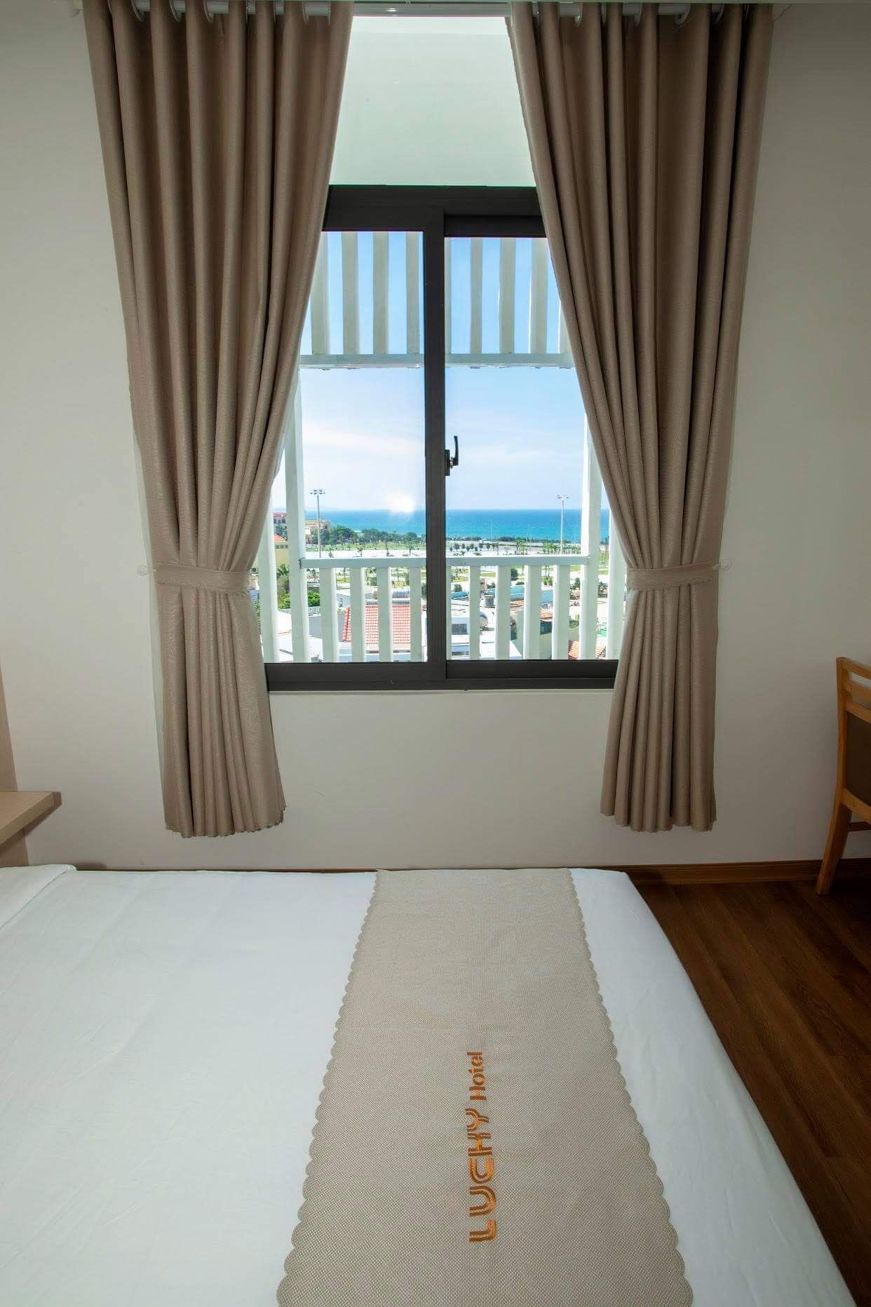 A bedroom with a large bed, beige curtains, and a view of a balcony and ocean.