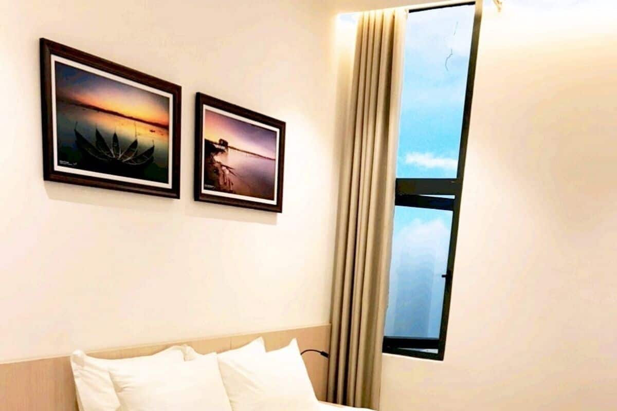 Een net opgemaakt bed met witte lakens, een beige plaid en een houten hoofdbord. Boven het bed hangen twee ingelijste foto’s en aan de rechterkant een raam met gordijnen.