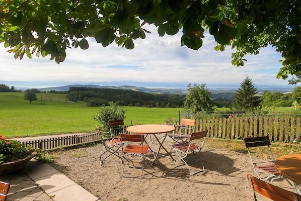 An outdoor patio with a round table and chairs, surrounded by a wooden fence, overlooking a green landscape with distant hills.