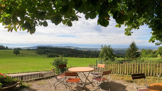 An outdoor patio with a round table and chairs, surrounded by a wooden fence, overlooking a green landscape with distant hills. - Family room Stammhaus category I - Höchsten Natur Erlebnis Hotel (Illmensee)