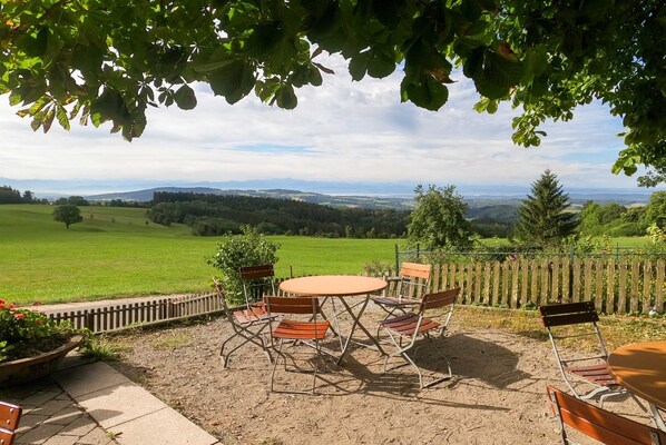 An outdoor patio with a round table and chairs, surrounded by a wooden fence, overlooking a green landscape with distant hills.
