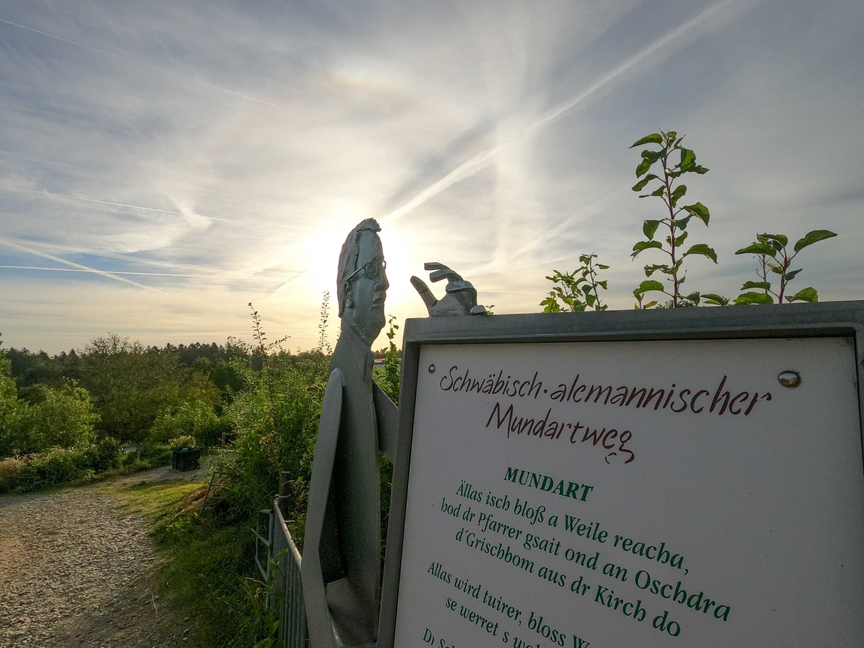 A sign with German text, a sculpture, and a view of a cloudy sky.