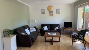 Een woonkamer met een donkerbruine bank, een witte kast, een houten salontafel en een rieten fauteuil. Er staat een televisie op een houten tafel en een lamp op de vloer.