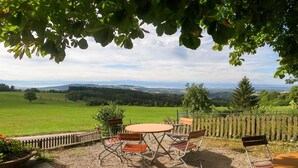 Une terrasse extérieure avec une table ronde et des chaises, entourée d’une clôture en bois, donnant sur un paysage verdoyant avec des collines à l’horizon.