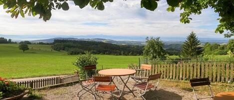 An outdoor patio with a round table and chairs, surrounded by a wooden fence, overlooking a green landscape with distant hills.