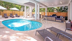 A covered outdoor swimming pool area with lounge chairs, a barbecue setup, and a wooden fence in the background.