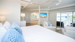 A modern hotel room with a large bed, a sitting area with a chair and a small table, a television, and a balcony with outdoor furniture.