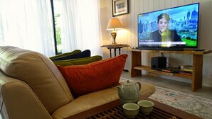 Una sala de estar con un sofá de cuero, almohadones de colores, una mesa de centro con una tetera y tazas, un televisor mostrando un noticiero, y una lámpara sobre una mesita auxiliar.