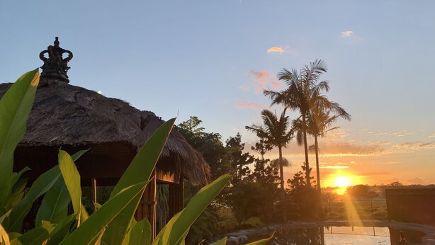 Una estructura con techo de paja y decoración en la parte superior, rodeada de follaje tropical y palmeras, con un atardecer de fondo.