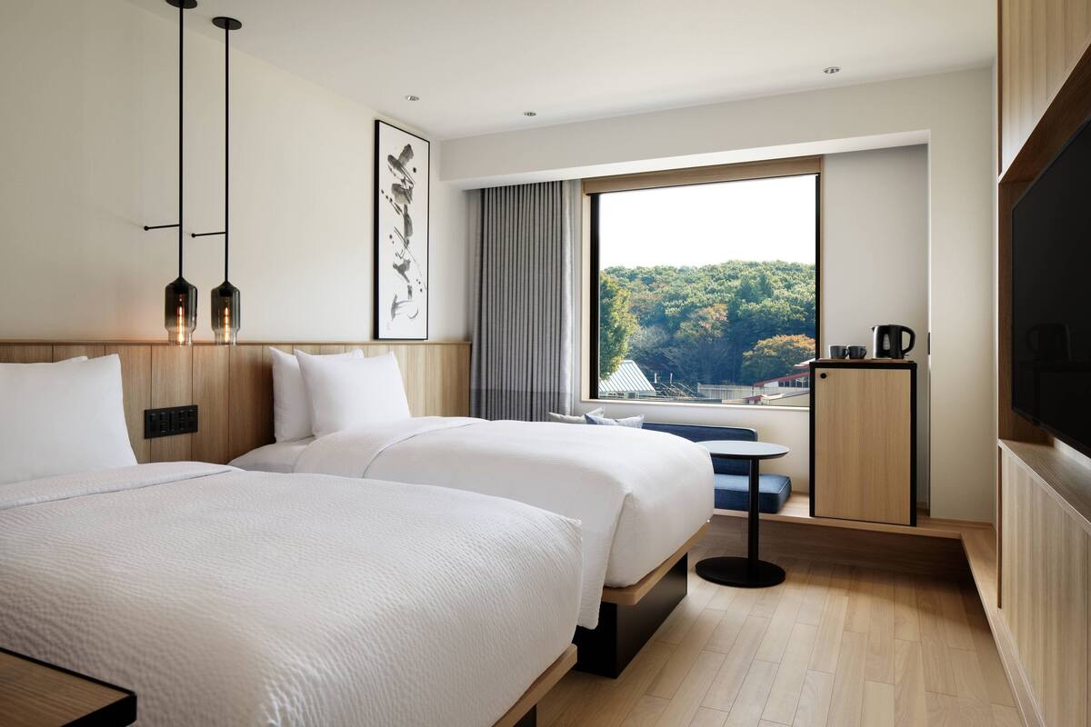 A modern hotel room with two beds, a large window with a view of a forest, a small table, and a TV.