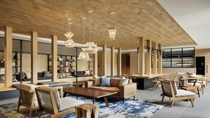 A spacious living area with a sofa set, wooden chairs, and a coffee table. There are hanging lights and a large window in the background.