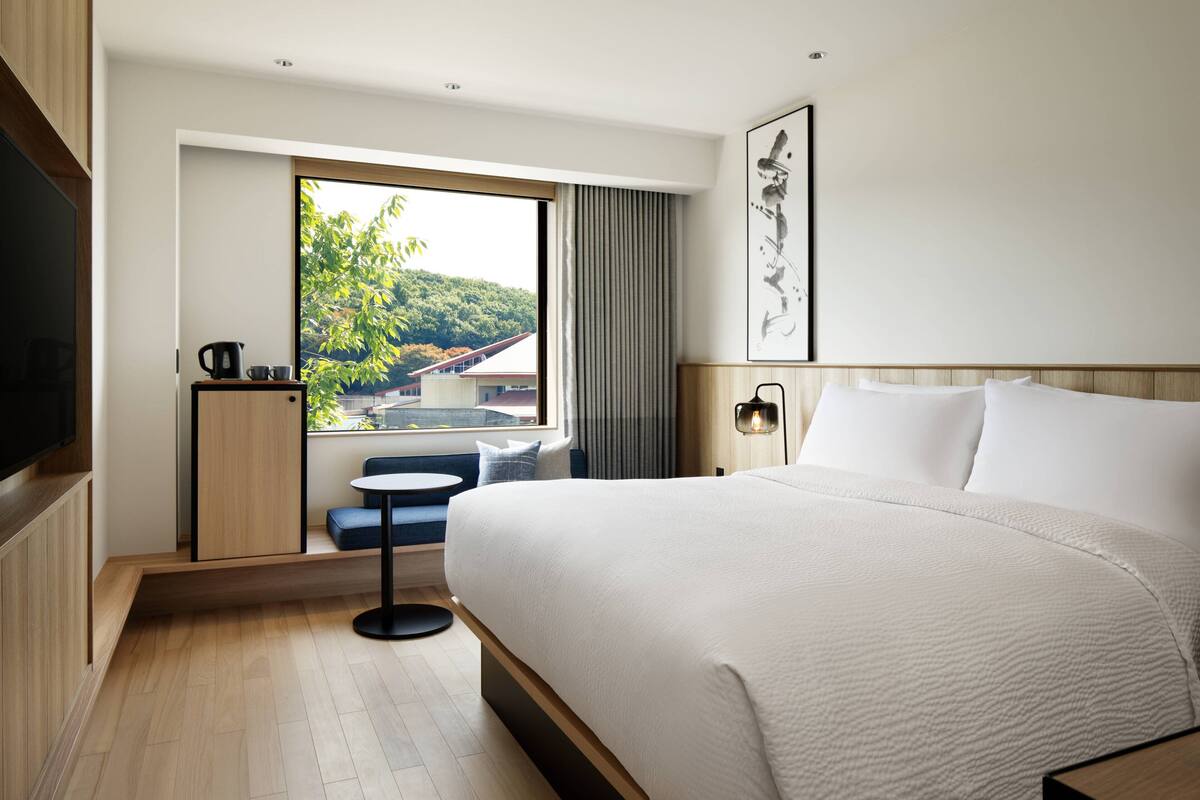 A modern hotel room with a large bed, a small table with a kettle, a seating area with a view of a green landscape, and a flat-screen TV.