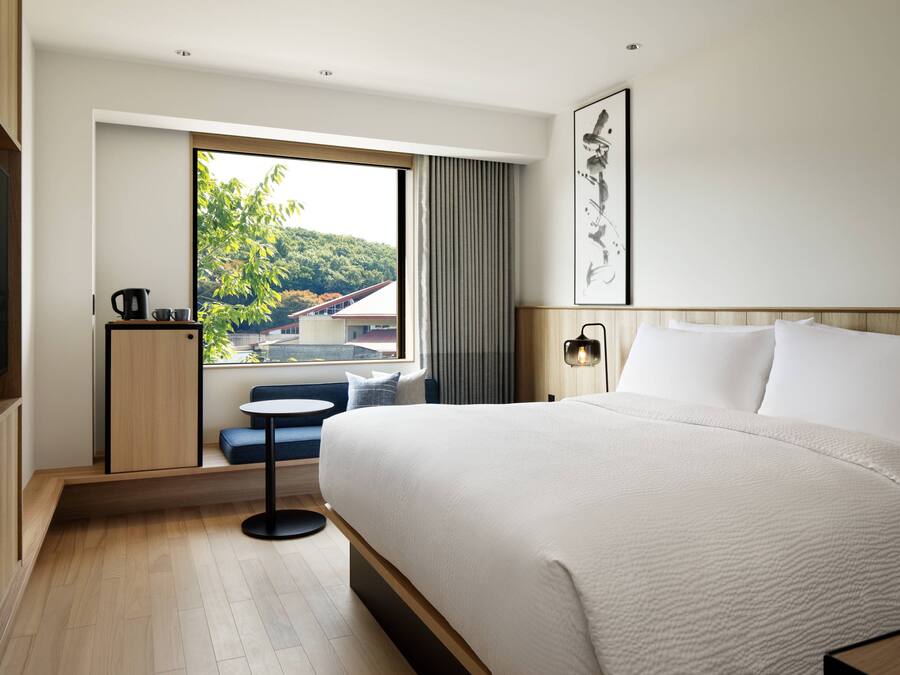 A modern hotel room with a large bed, a small table with a kettle, a seating area with a view of a green landscape, and a flat-screen TV.