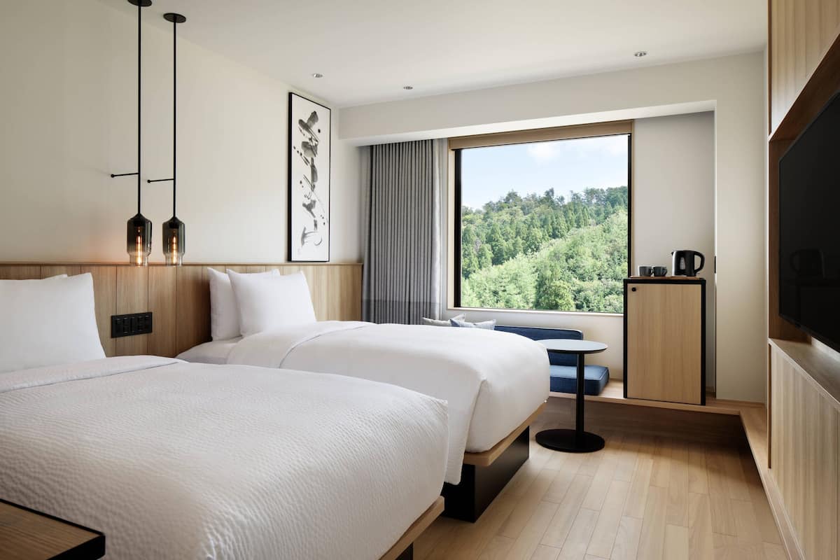 A modern hotel room with two beds, a large window with a forest view, a small table, and a TV.