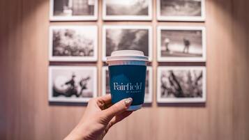 A hand holding a Fairfield by Marriott coffee cup in front of a wall with framed black and white photographs.