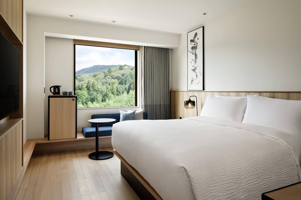 A modern hotel room with a large bed, a small seating area, a window with a view of greenery, and a flat-screen TV.